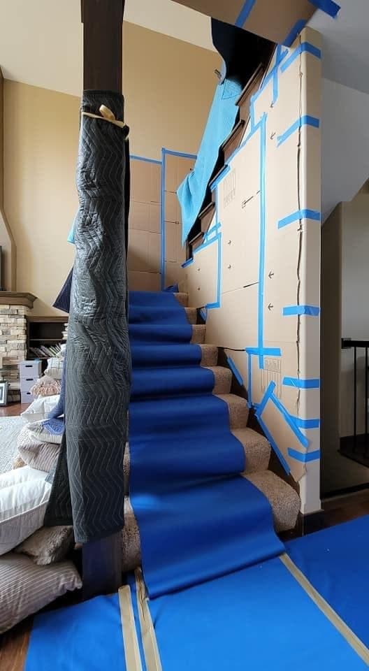 A staircase with a blue runner down the treads, cardboard taped to the wall and the newel post wrapped in a moving blanket